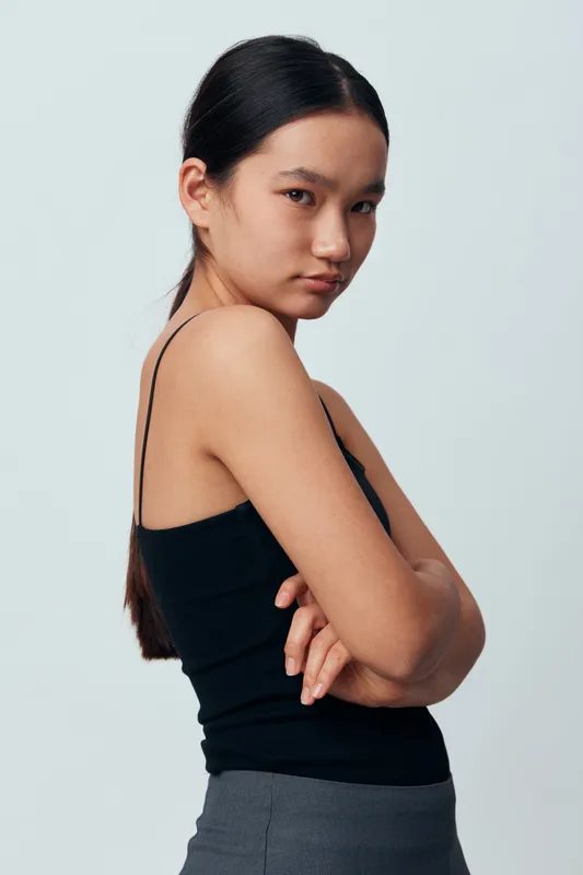 Young woman with long dark hair wearing black sleeveless top and gray pants posing sideways against plain light background with confident expression and crossed arms.