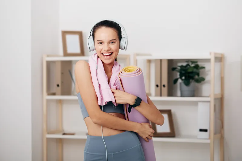 Active woman fitness enthusiast wearing headphones, smiling in a bright home gym, holding a yoga mat and towel, ready for a workout session and motivation.