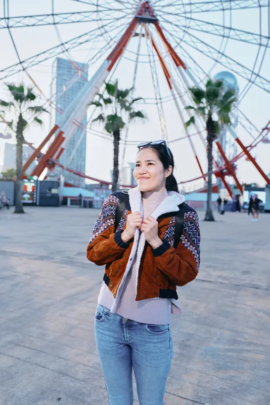 Woman wearing a brown jacket and scarf smiles near a ferris wheel in an urban plaza, palm trees and modern buildings visible, sunny outdoor scene conveying travel and casual city life.