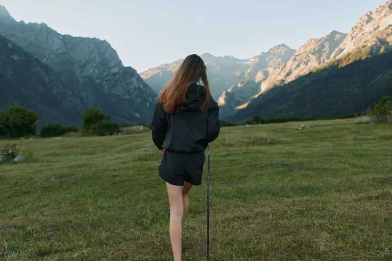 A lone hiker stands in a grassy valley with towering mountains in the distance. She wears a dark jacket and shorts, holding a trekking pole, enjoying the scenic outdoors.