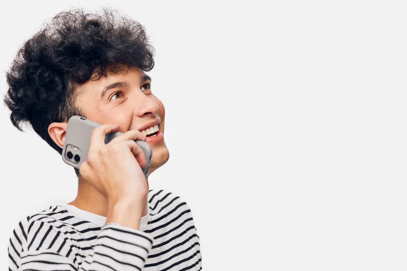 smiling man talking on smartphone wearing striped shirt with curly hair on white background, cheerful and casual communication concept