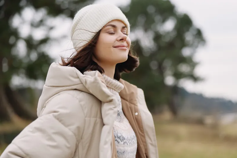Woman in a beige winter coat and knit cap stands outdoors with closed eyes and a serene smile, natural light, soft breeze and blurred trees in the background.