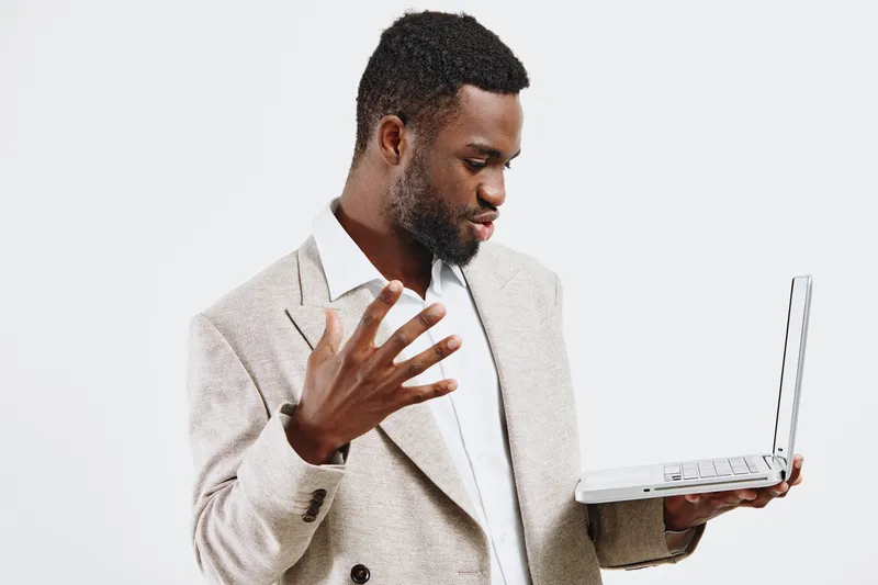 Businessman with dark skin reading and gesturing while looking at his laptop, dressed in a beige blazer and white shirt, isolated on a plain white background. Office, technology, work, professional co