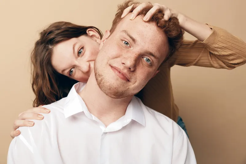 Young couple with playful mood, woman hugging man from behind and touching his hair. Studio portrait on beige background, concept of love, friendship, and happiness.