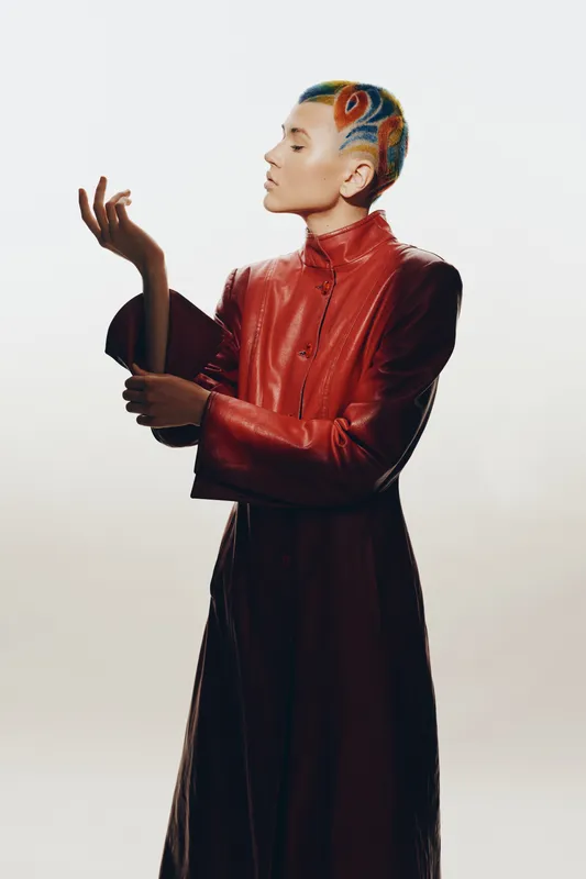 Woman with colorful shaved hair wearing a red leather coat, posing thoughtfully against a plain light background with dramatic lighting and minimalist style.