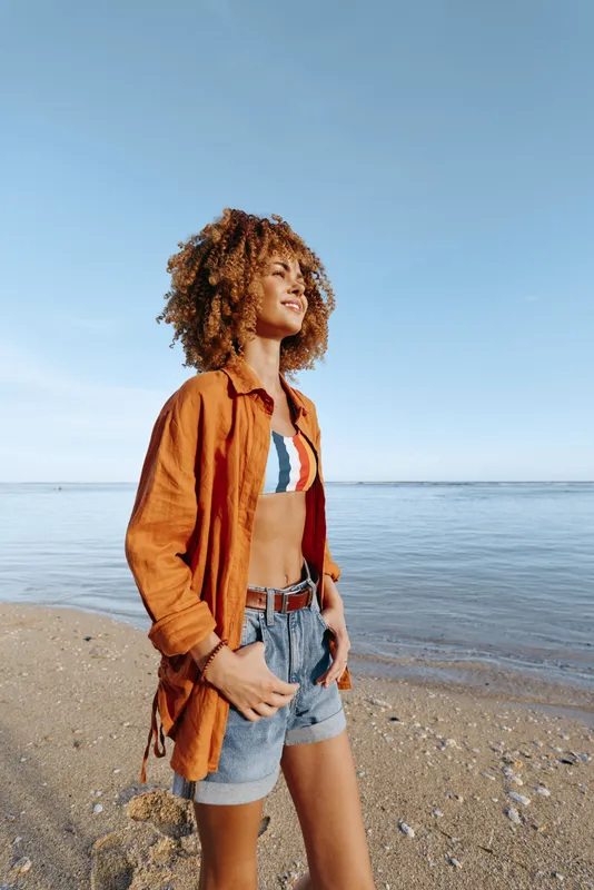 Smiling woman on the beach wearing an orange shirt, denim shorts, and striped bikini top.