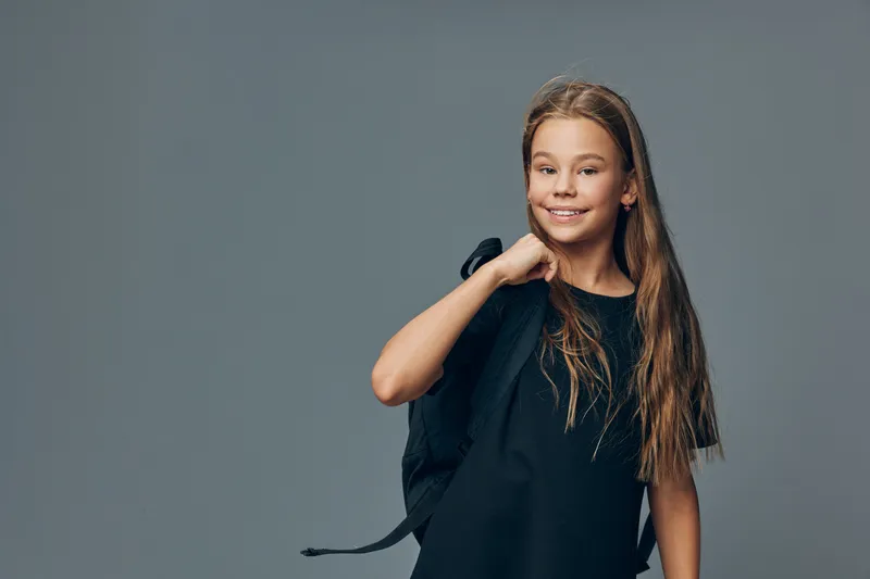 Girl with long hair wearing black dress holding backpack on shoulder smiling and looking at camera against gray studio background. Child posing with confidence and casual style.