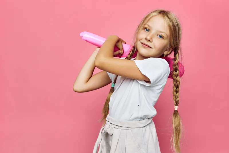 Smiling girl with long braids holding pink toy bat, posing confidently on a solid pink background. Studio shot concept for childhood fun.