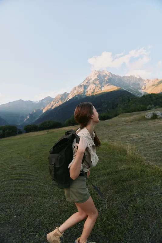 A young woman with a backpack hiking in the mountains during daytime looks up at the snow-capped peak. The scene captures outdoor adventure and active lifestyle.