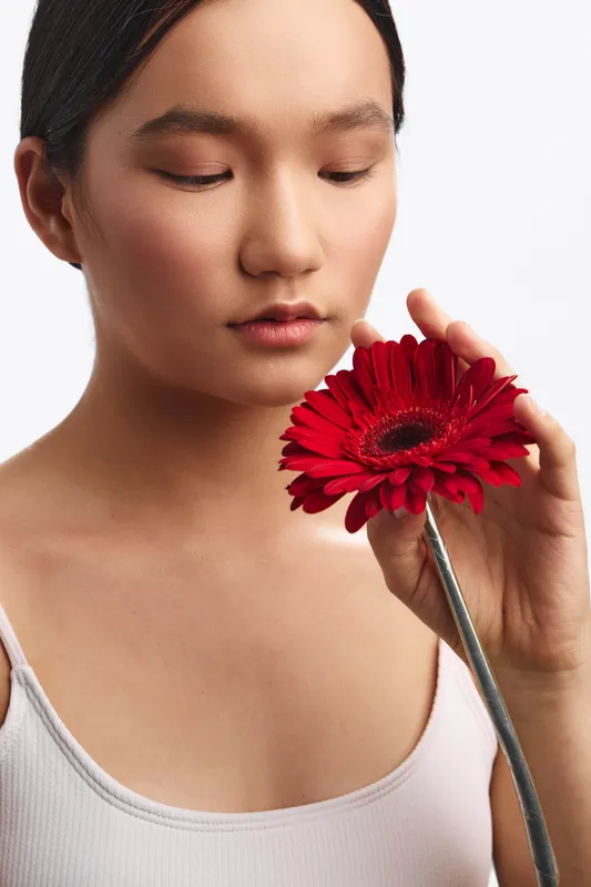 Asian woman holds a vibrant red flower near her face in a studio setting, her expression serene and focused, creating an intimate beauty moment.