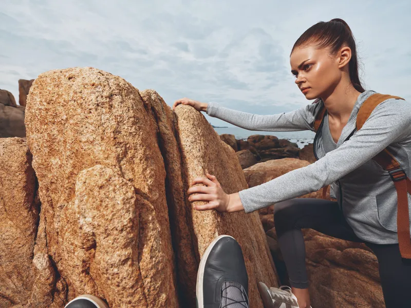 Young woman climbing on rocky terrain, showcasing determination in a gray athletic outfit with a backpack against a cloudy sky backdrop