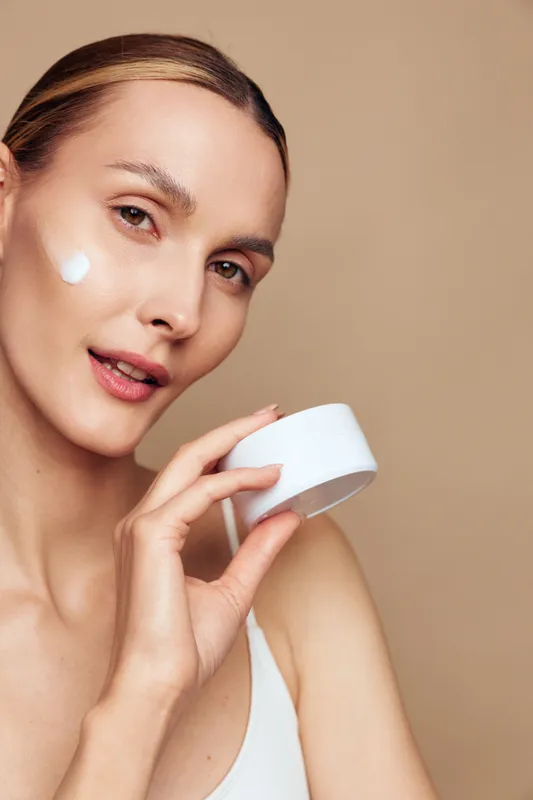 Woman skincare face cream selfcare beauty closeup wellness, young adult applying white moisturizer from a small jar, smooth skin, warm studio lighting, calm selfcare concept on beige background