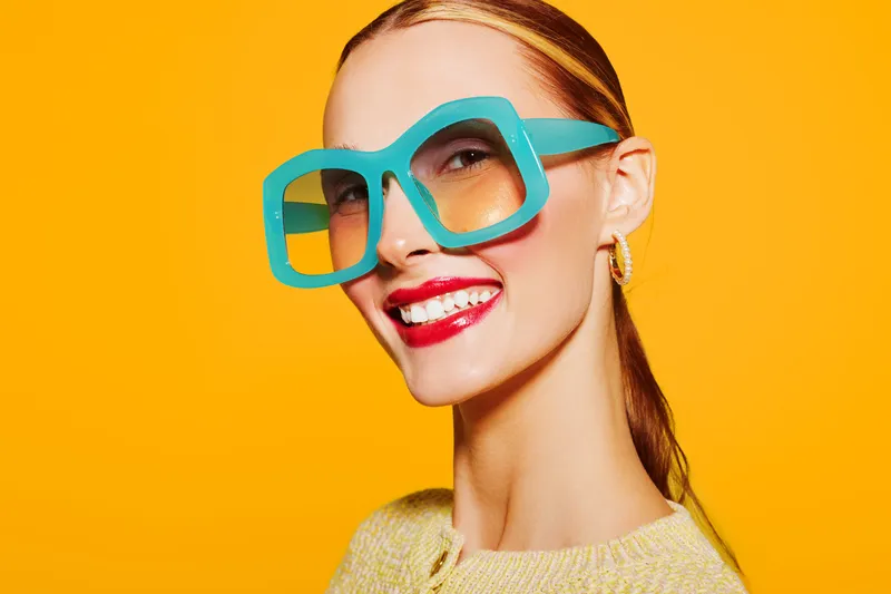 Smiling woman with bright red lipstick wears oversized turquoise glasses and yellow sweater against vibrant orange background. Concept of fashion, style, beauty, and confident expression.
