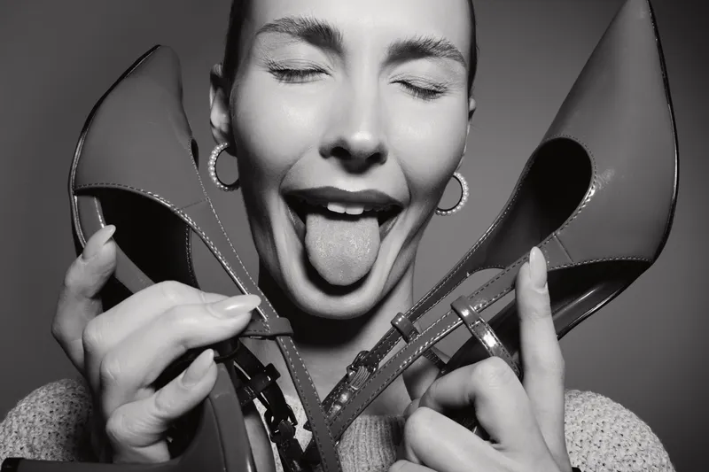 Young Caucasian woman with light skin plays with red high heels, smiling with closed eyes and tongue out, studio portrait in black and white.