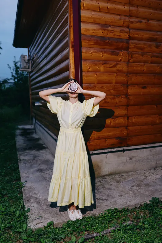 A woman in a yellow dress stands outdoors beside a wooden cabin at dusk, shielding her eyes with hands, creating a calm rustic fashion moment for storytelling