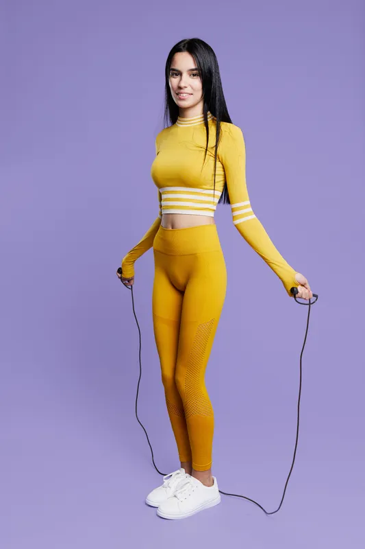 Woman sport fitness in yellow athletic wear holds jump rope, standing confidently on purple background, showcasing healthy lifestyle and active workout routine.