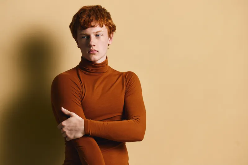 Young man with red hair in brown turtleneck sweater posing with folded arms against beige background. Studio portrait of thoughtful male with fair skin and casual outfit in minimal setting.