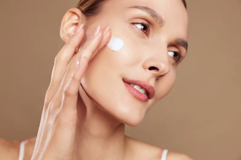 Smiling woman applying face cream with fingertips, closeup on warm beige background, beauty skincare routine, wellness and self care concept, soft studio lighting, calm mood
