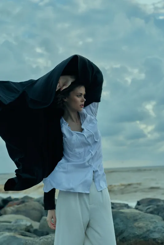 Woman stands on a rocky coast, wind lifting a flowing cloak as she gazes toward the ocean, white shirt and light pants beneath, moody sky, cinematic fashion editorial with dramatic mood