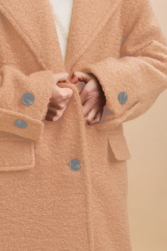 Asian woman is shown wearing a soft textured coat, standing in a neutral setting, her hands fastening the large buttons, conveying elegance, confidence, and timeless style.