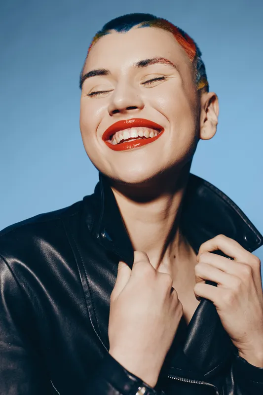 Smiling woman with colorful hair and bright red lipstick wearing a black leather jacket, expressing joy and confidence against a clear blue background.