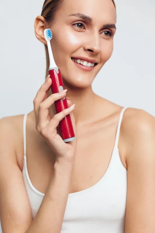 Young woman with clear skin holds red electric toothbrush near face smiling gently in white tank top isolated on light background, showing daily dental hygiene routine and positive mood