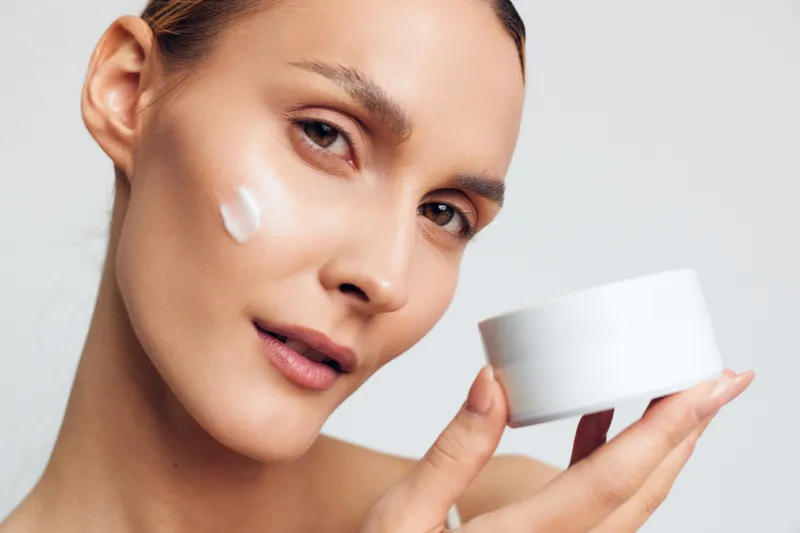 Young woman applying white face cream on cheek holding a cosmetic jar. Close-up portrait with smooth skin on light background. Concept of skincare beauty health and self-care.