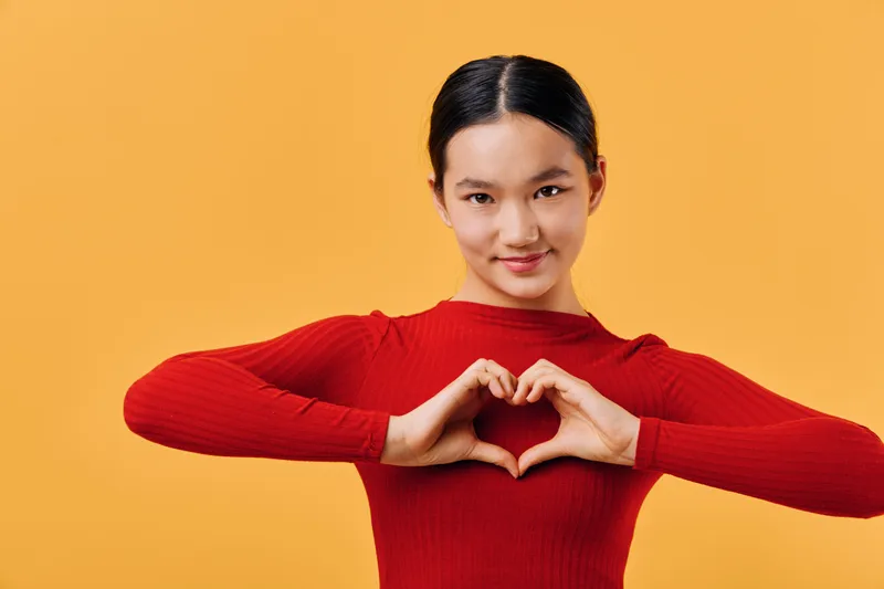 Young woman with dark hair and red sweater making heart shape with hands in front of yellow background. Smiling female showing love and positive gesture with fingers.