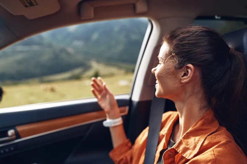Woman seated in a car, gazing out the window at a scenic landscape, enjoying a peaceful travel moment under bright daytime light in a moving road setting.
