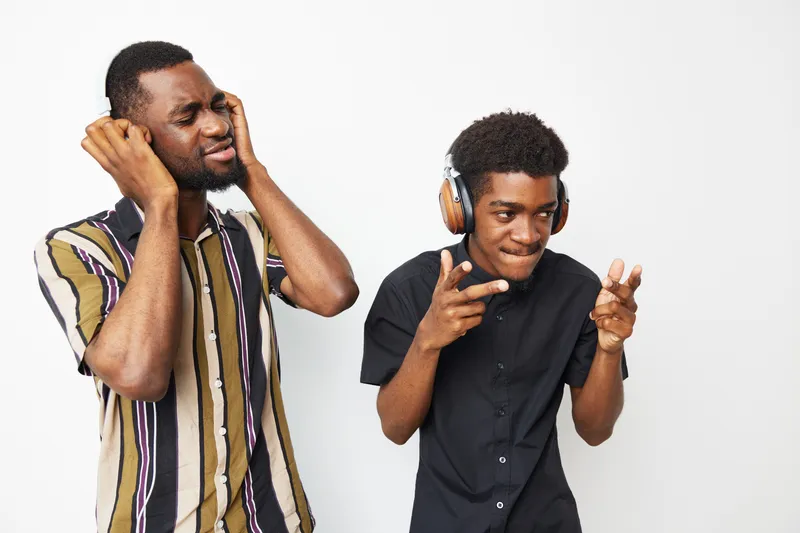 Two black men enjoying music together, one smiling while the other reacts dramatically. The joyful atmosphere captures friendship and fun moments.