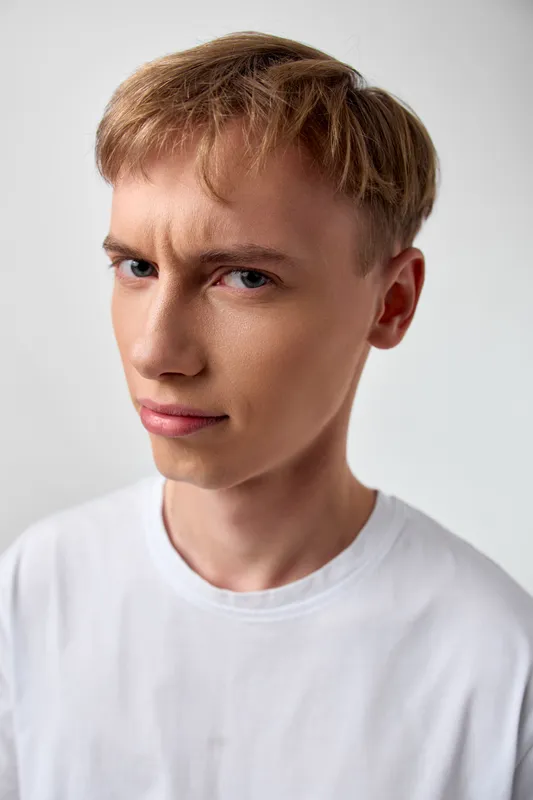 Close up of a young man with a confident expression, wearing a white shirt under soft studio lighting, neutral background, suitable for fashion, lifestyle, and headshot style ideas