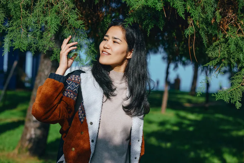 Woman in a brown shearling jacket stands outdoors among green trees, sunlight filters through leaves, casual fashion, relaxed pose, cheerful mood, natural park setting, candid lifestyle photograph
