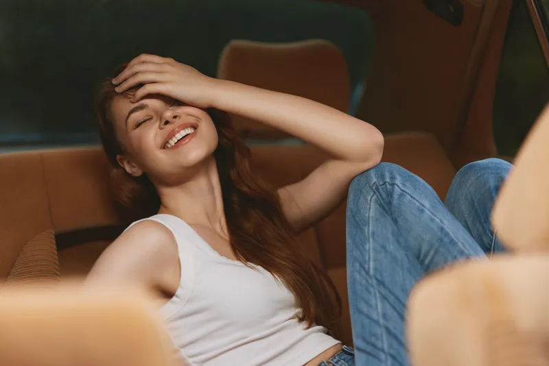 Cheerful young woman enjoying relaxation in a cozy car interior, wearing casual attire and exuding happiness with natural light and warm tones.