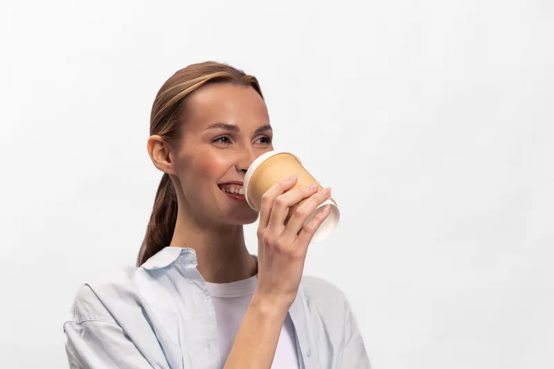 Smiling young woman with brown hair drinks coffee from a disposable cup. Casual outfit and natural makeup. Concept of refreshment, morning routine, and relaxation.