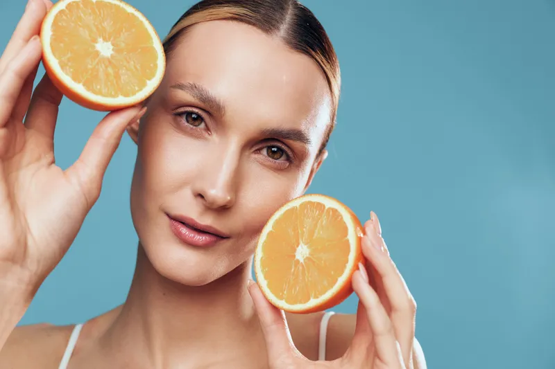 A young woman with clear skin holds two halves of a fresh orange near her face against a blue background. Concept of natural beauty, skincare, and healthy lifestyle.