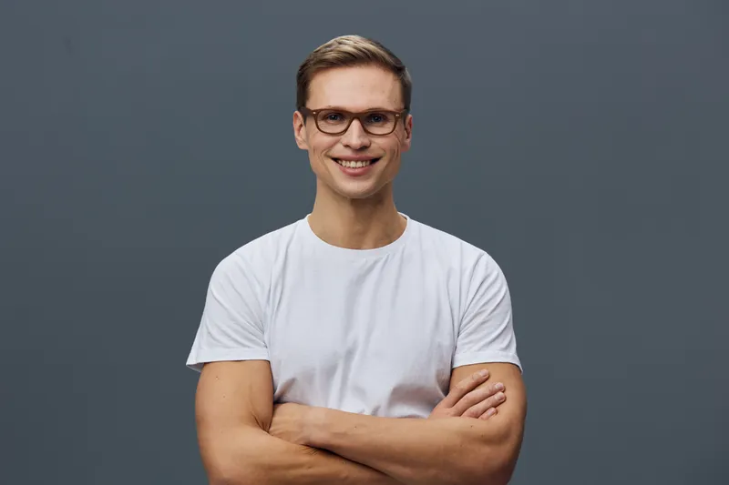 Smiling young caucasian man wearing glasses and a plain white T-shirt, looking confidently at camera with arms crossed, isolated on a solid gray background. Studio portrait for lifestyle and happiness