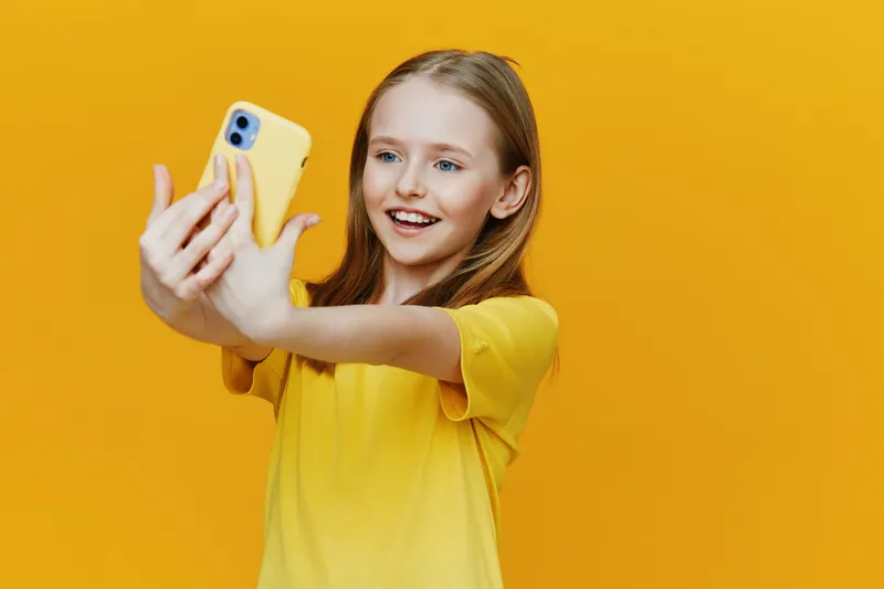 Happy girl taking a selfie with a yellow smartphone against a bright yellow background, showcasing joy and youthful energy in a vibrant summer setting