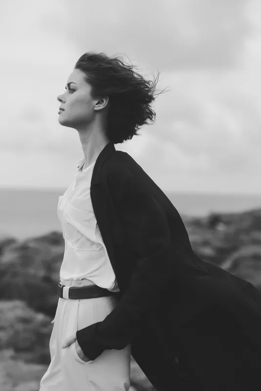 Black and white silhouette of a woman on a windy coast, eyes closed, coat billowing, moody mood, solitary reflection by the sea, minimalist fashion vibe and sense of freedom against rugged shoreline