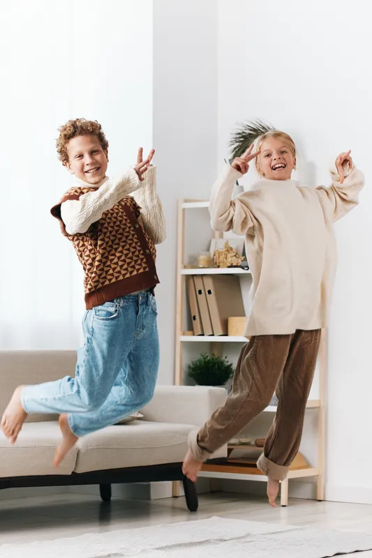 Two cheerful children jump on a light sofa indoors, smiling and carefree, capturing playful energy and lively motion in a bright, cozy home setting.