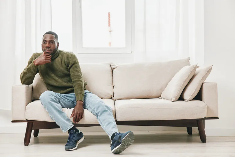 Casual portrait of a Black man sitting on a modern sofa, wearing a green sweater and jeans, expressing contemplation against a bright interior backdrop