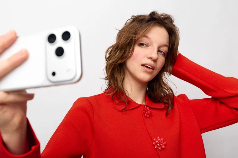 Young woman in red sweater takes selfie with smartphone on white background. Casual style, natural makeup, and relaxed expression. Concept of social media and self-portrait.