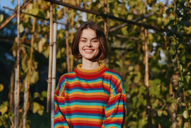 Woman smiling in a colorful striped sweater, young adult female model standing in a sunlit garden trellis, casual outfit, short hair, relaxed portrait for lifestyle and fashion.