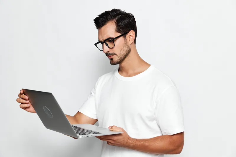 Thoughtful man in stylish glasses holding laptop, looking at screen with concentration. Isolated on plain solid background, capturing focus and curiosity.