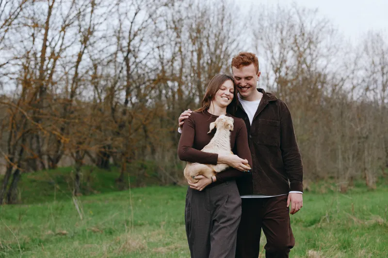 Joyful couple outdoors with a small dog, embracing and smiling, casual outfits, warm mood, green field and bare trees in the background, candid lifestyle moment