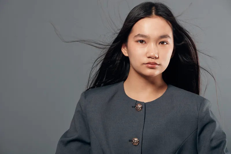 Young woman with long black hair wearing gray jacket posing confidently against plain gray background for fashion portrait studio photo session. Serious expression and natural look.
