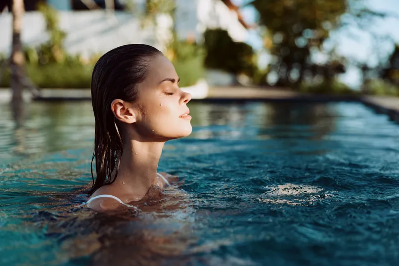 Young woman with wet hair relaxing in outdoor pool water under sunlight. Lifestyle concept of leisure, wellness, and summer vacation. Calm expression with natural background blurred.