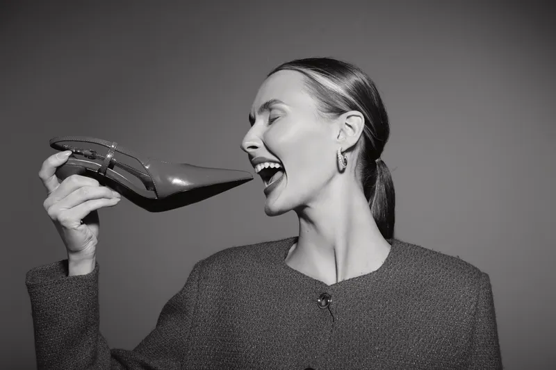 Caucasian woman with light skin laughs holding a high heel shoe near her face in studio. Concept of fashion and joy.