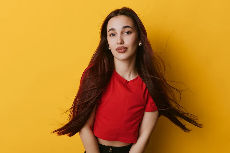 Young woman with long hair wearing red shirt posing against yellow background, looking at camera with confident expression and natural makeup in studio portrait.
