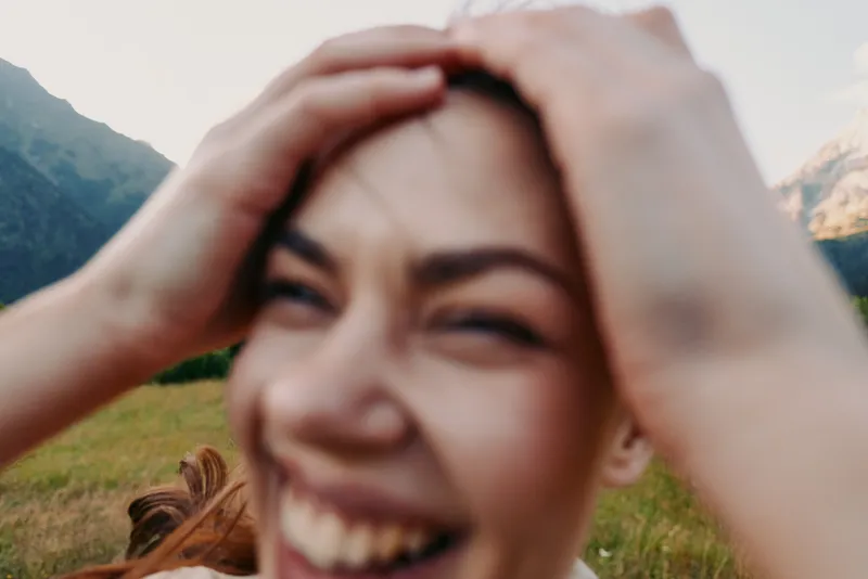 Young woman with long hair laughs joyfully while holding her head outdoors in a natural mountainous landscape during daytime.