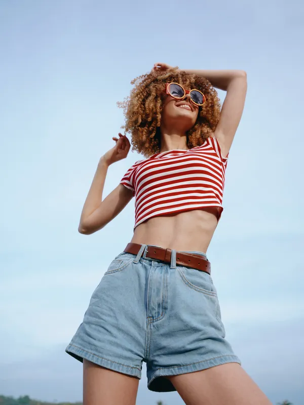 A young woman stands outdoors in a striped crop top and high waisted denim shorts, posing confidently against a clear blue sky, sunny and vibrant.